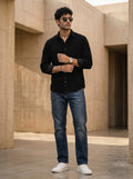 Man wearing a black shirt, blue jeans, and white sneakers standing in front of beige architectural structures.